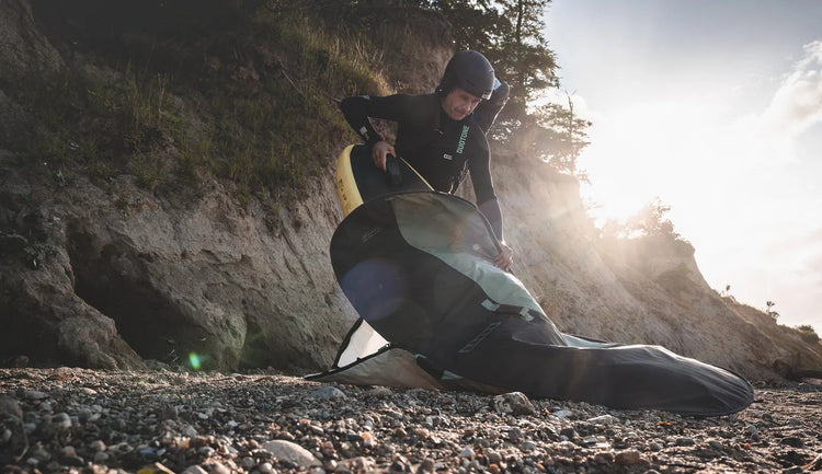 Green Hat Kiteboarding-Collection-Clearance Wingfoiling Bags-Man Putting a Wing Board in an Ion Foilboard Bag on a Rocky Beach