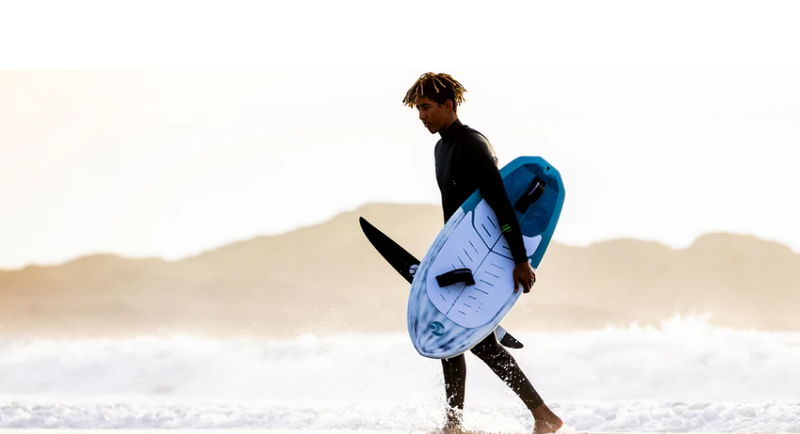 Load image into Gallery viewer, Green Hat Kiteboarding-2024 Cabrinha 04 Link Wing Board-Man in a wetsuit carrying a Link Board on a beach with sand dunes in the background