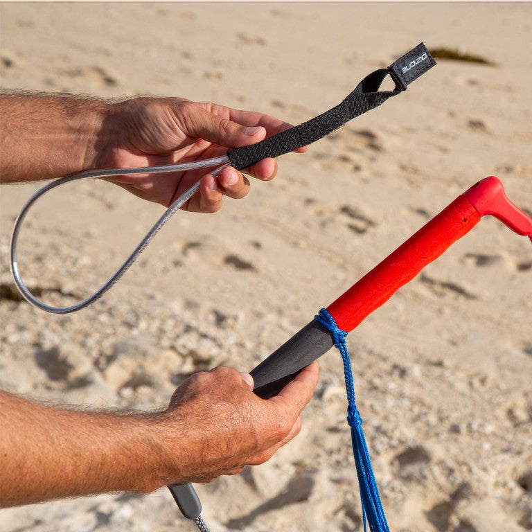 Load image into Gallery viewer, Green Hat Kiteboarding-Ozone Wing/Parawing Harness Line-Man holding a parawing bar in one hand and a harness line in the other against a sandy background