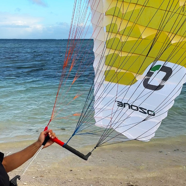 Load image into Gallery viewer, Green Hat Kiteboarding-Ozone Wing/Parawing Harness Line-Man holding a parawing in one hand hooked in the harness line on a beach with ocean in the background