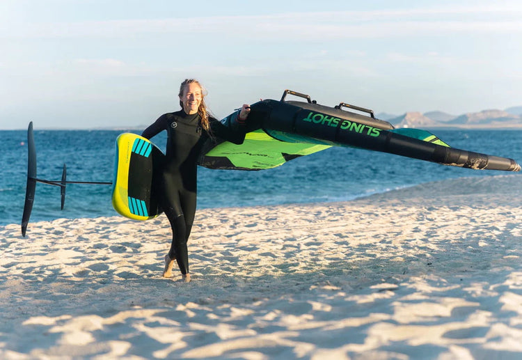 Green Hat Kiteboarding-Collection Recent Price Drop Wing Foiling-Woman Wearing a Wetsuit Carrying Wing Foil Gear on a Beach in Baja