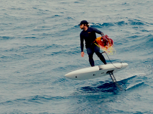 Green Hat Kiteboarding-F-One FRIGATE Parawing-Man in a wetsuit riding a hydrofoil board on water with a parawing stashed in his hand