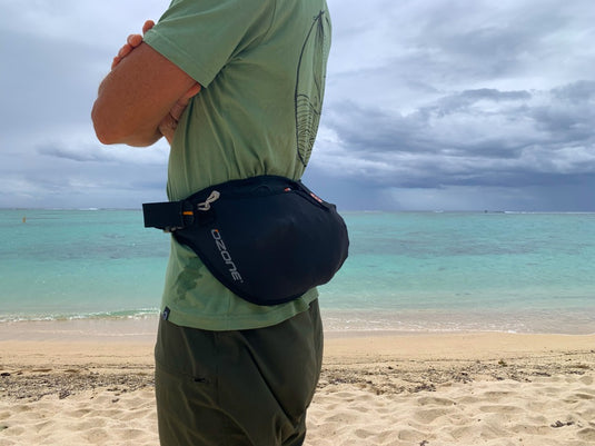 Person standing on a beach with a Ozone Parawing Stash Harness, facing the ocean under a cloudy sky.